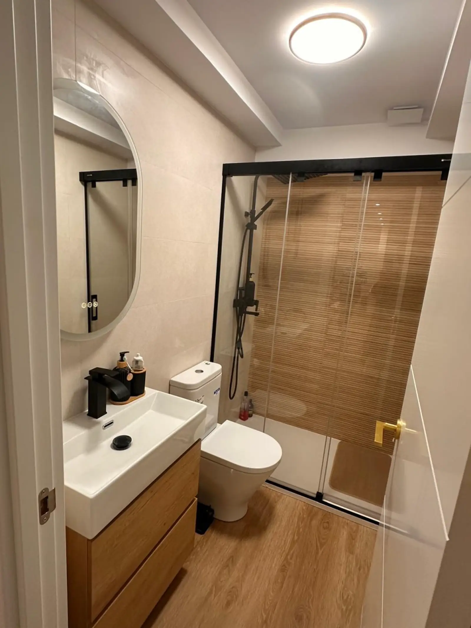 Baño reformado con ducha, mampara negra, mueble de madera y espejo redondo